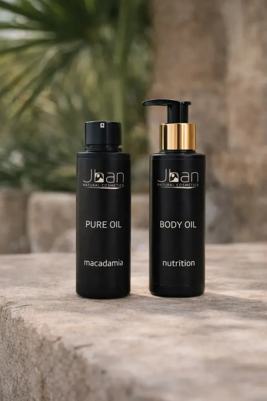 Косметика Jhan Natural Cosmetics на металлическом подносе: масла для тела, волос и лица, рядом белые цветы на текстурном светлом фоне.